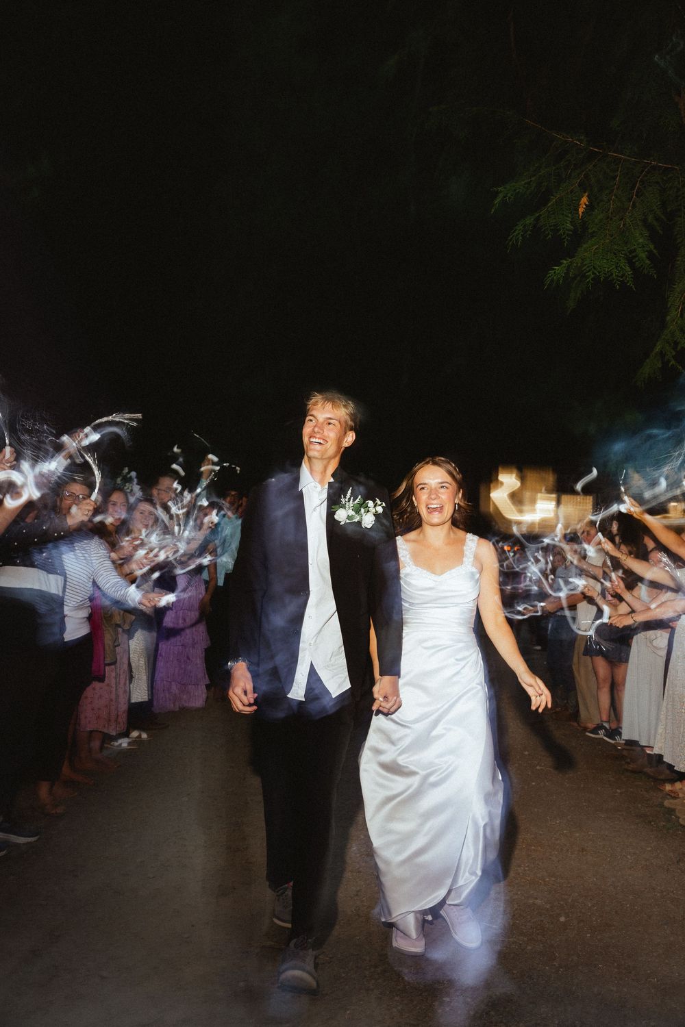 Bride and groom walk hand-in-hand through a sparkler exit tunnel at their outdoor night wedding celebration.