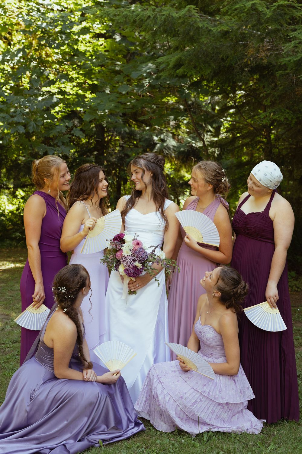 Bride in white gown with bridesmaids in purple dresses holding white fans, posing outdoors surrounded by lush green trees.