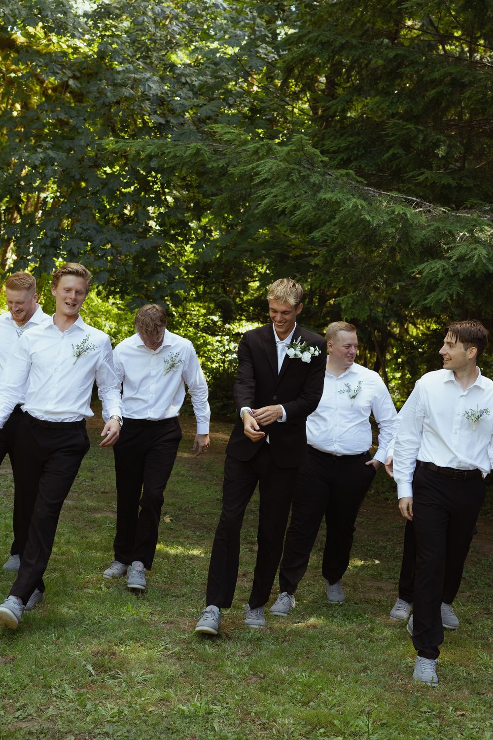 Groom in black suit walks with five groomsmen in white shirts through a sunny green forest setting.