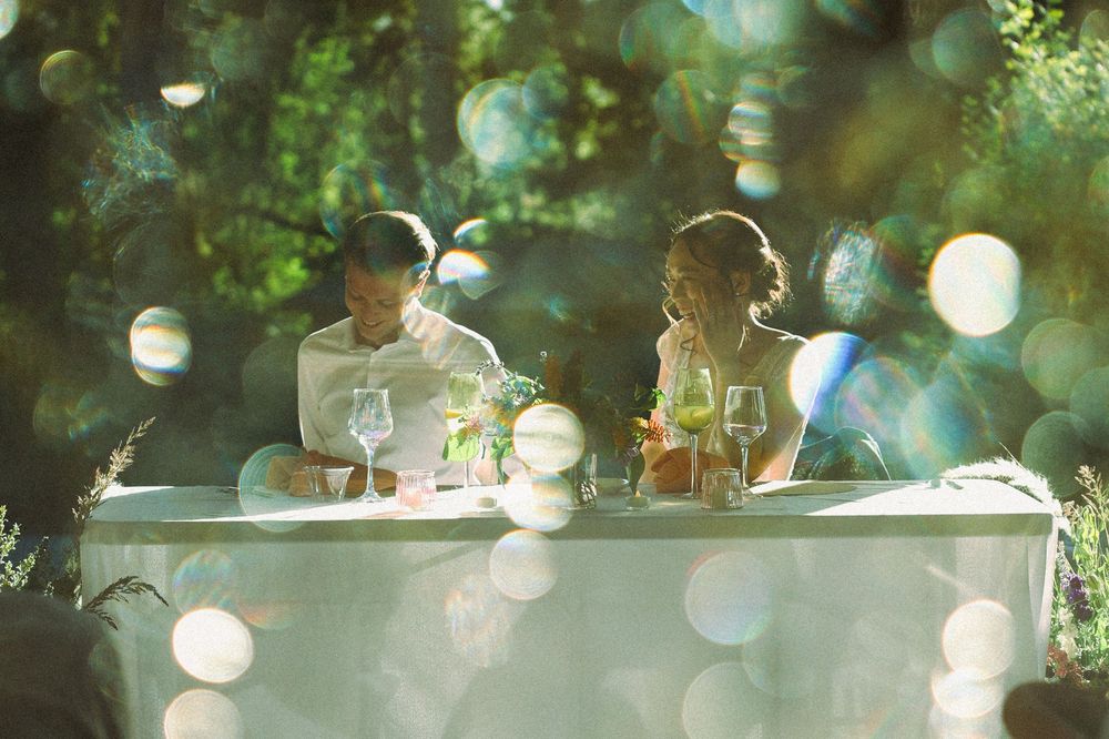 Couple dining outdoors at elegant table surrounded by lush green trees with dreamy bokeh light effects.