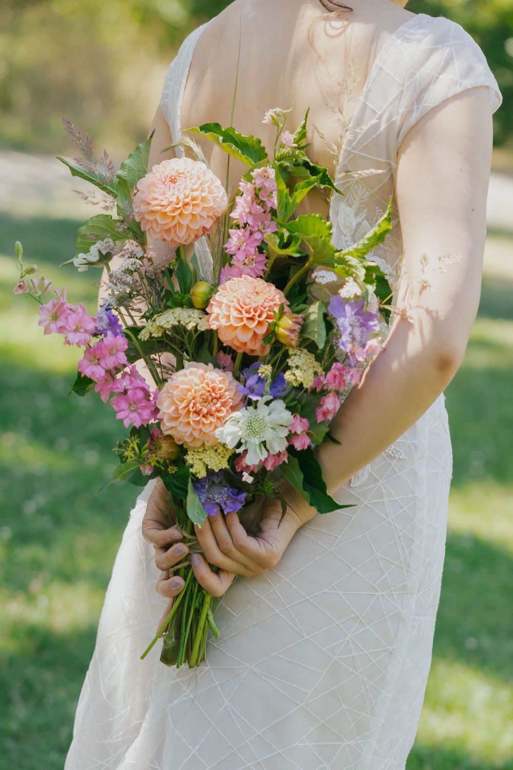 Bride in white lace dress holding a lush bouquet of peach dahlias, pink, and purple wildflowers outdoors.