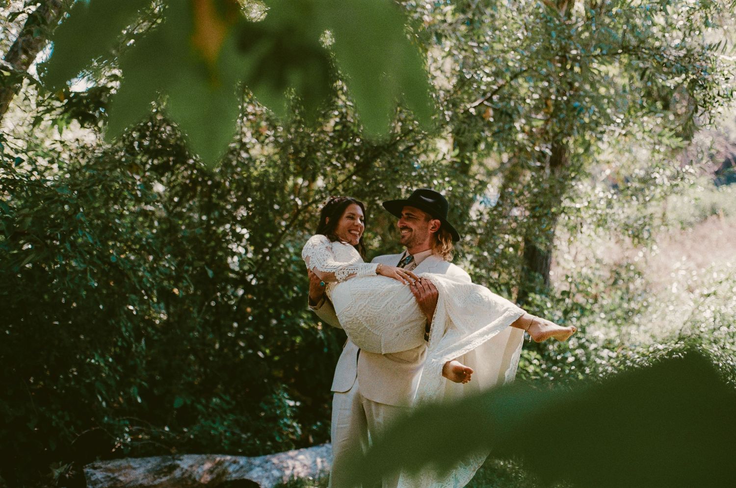 Groom lifting laughing bride in lush green garden setting during outdoor wedding photo session.