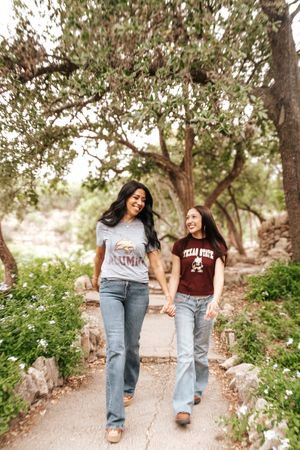 Two friends in casual outfits walking together on a scenic path lined with trees and wildflowers on a sunny day.