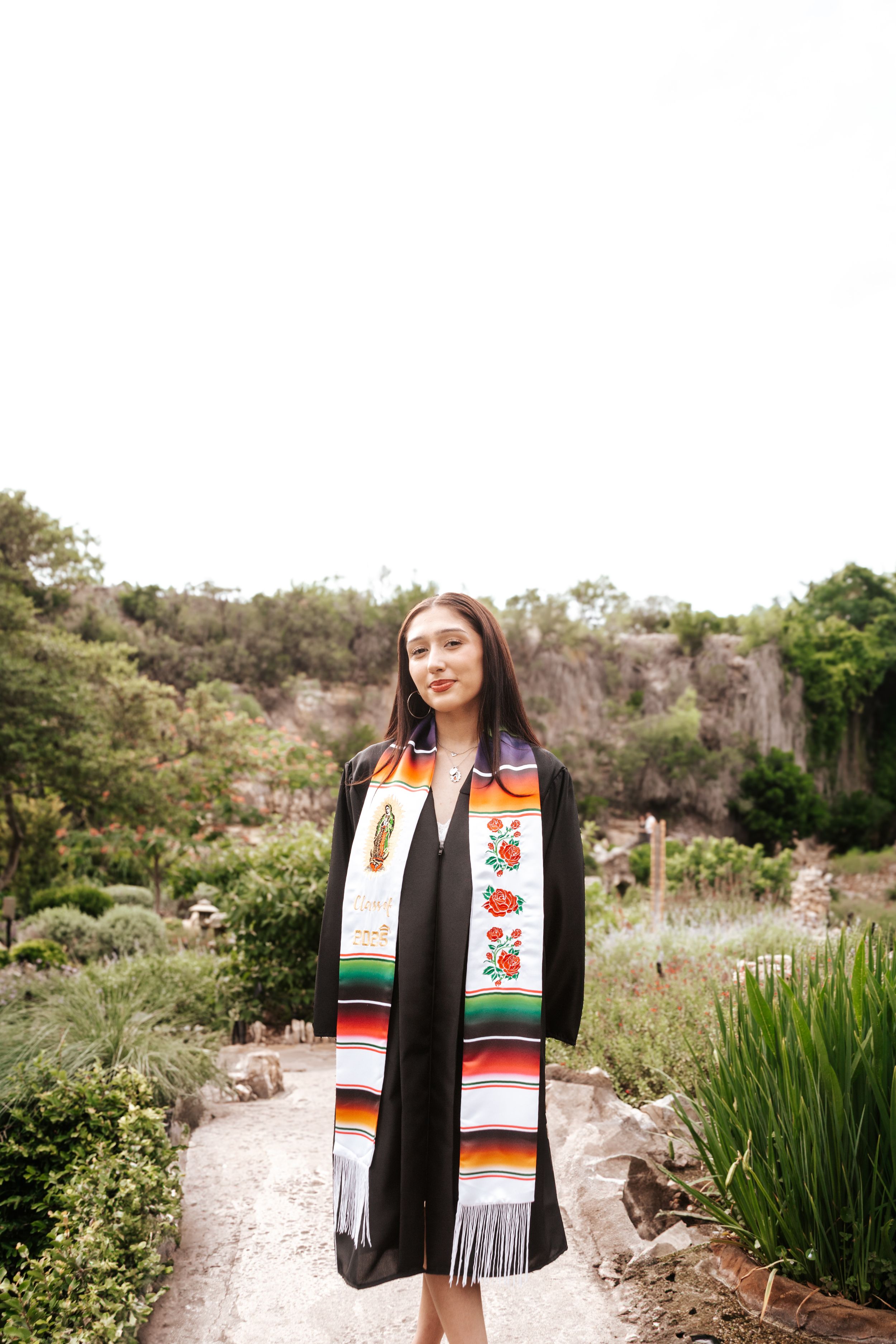 Graduate wearing cultural stole and black graduation gown poses on garden path with rocky cliffs in background.