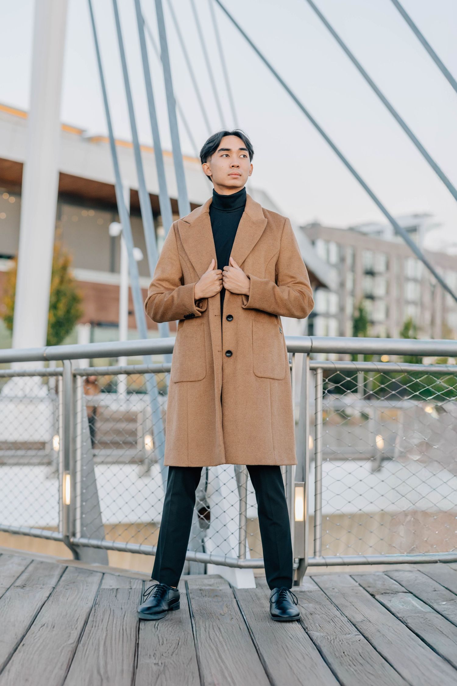Stylish young Asian man in tan overcoat standing on pedestrian bridge with cable supports behind him.