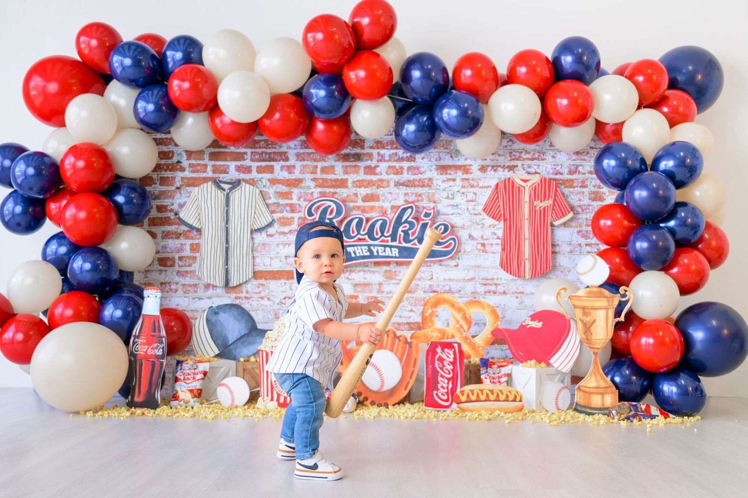 Festive red, white and blue balloon arch decorates a brick wall backdrop for a baseball-themed celebration setting.