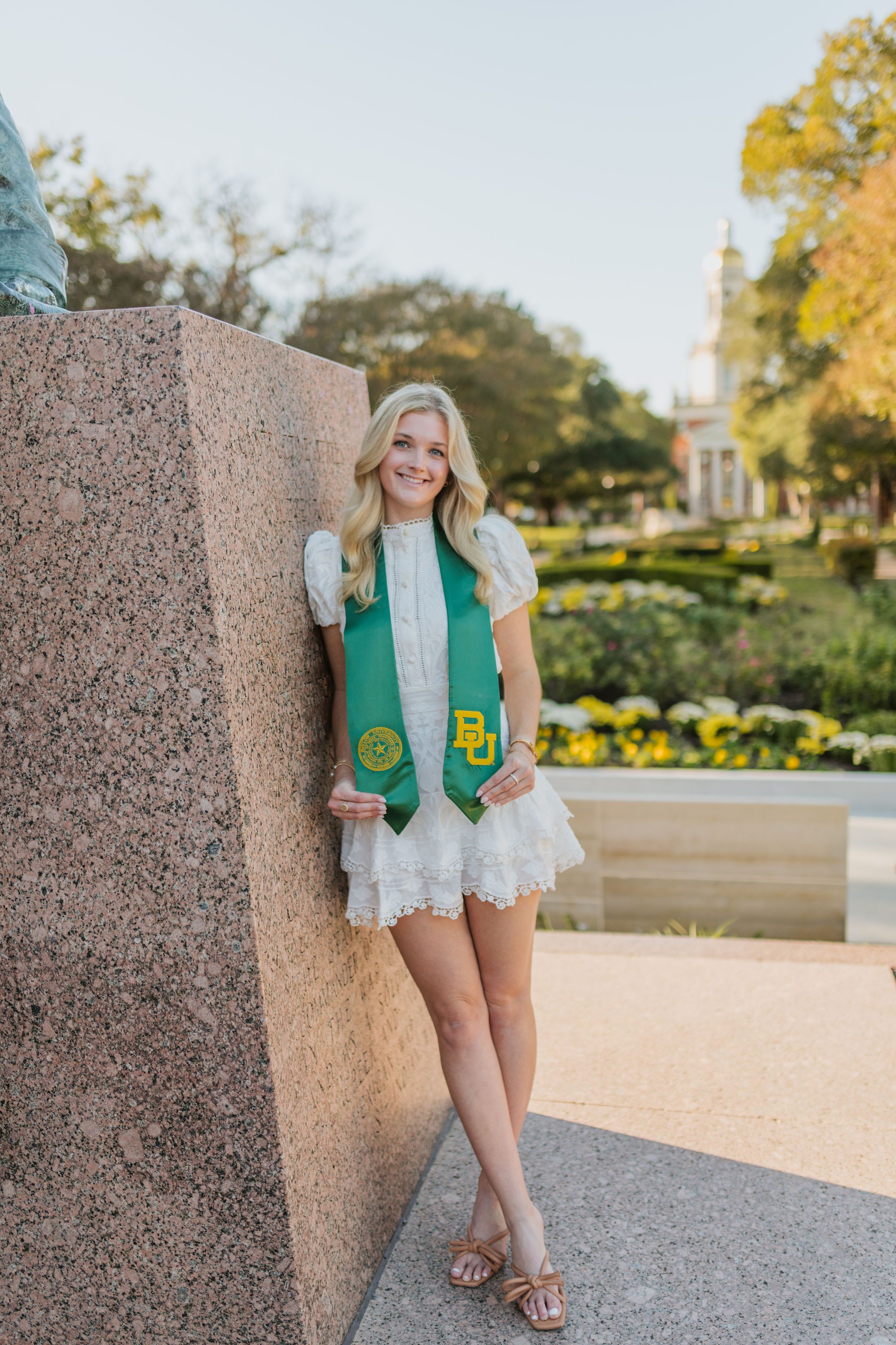 Catherine Kramer, smiling blonde woman in a white dress with a green Baylor BU graduation stole, leaning against a stone monument.