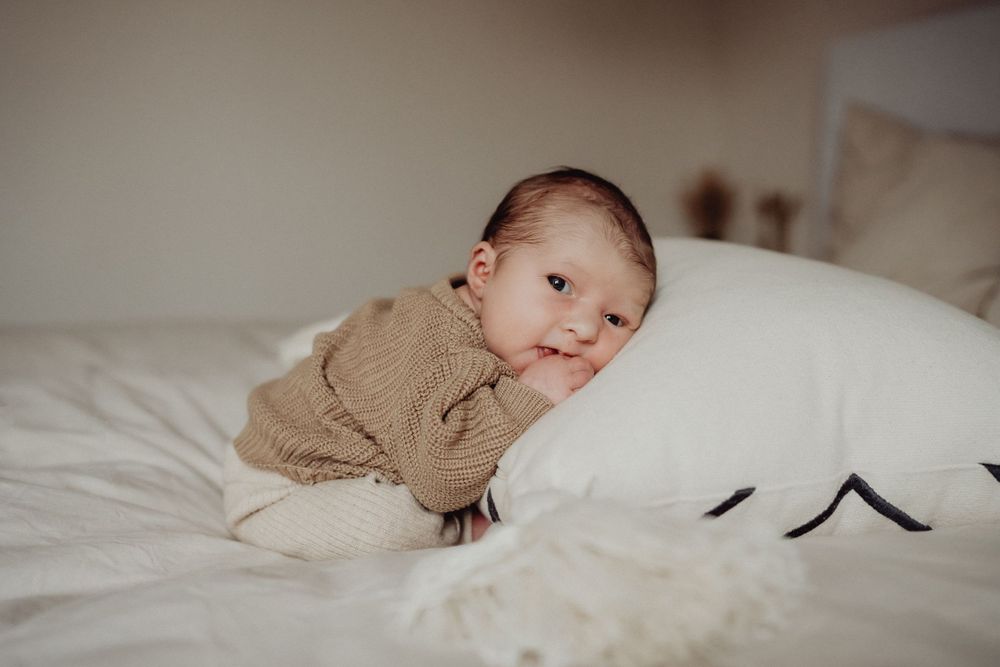 Baby in brown sweater resting on white bedding with stuffed animal.