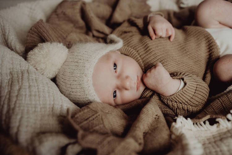 Infant wearing cozy brown sweater lying on soft textured bedding in natural light.