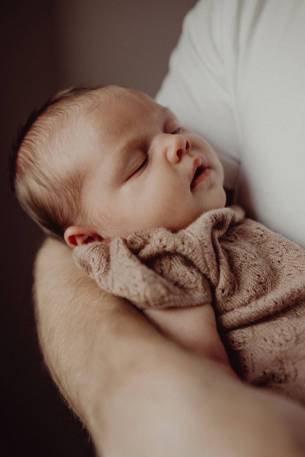 Sleeping infant wrapped in beige blanket being cradled in gentle lighting.