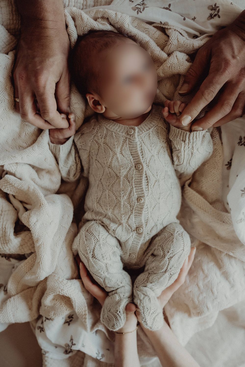 Infant wearing knit onesie lying on textured blanket with caring hands nearby.