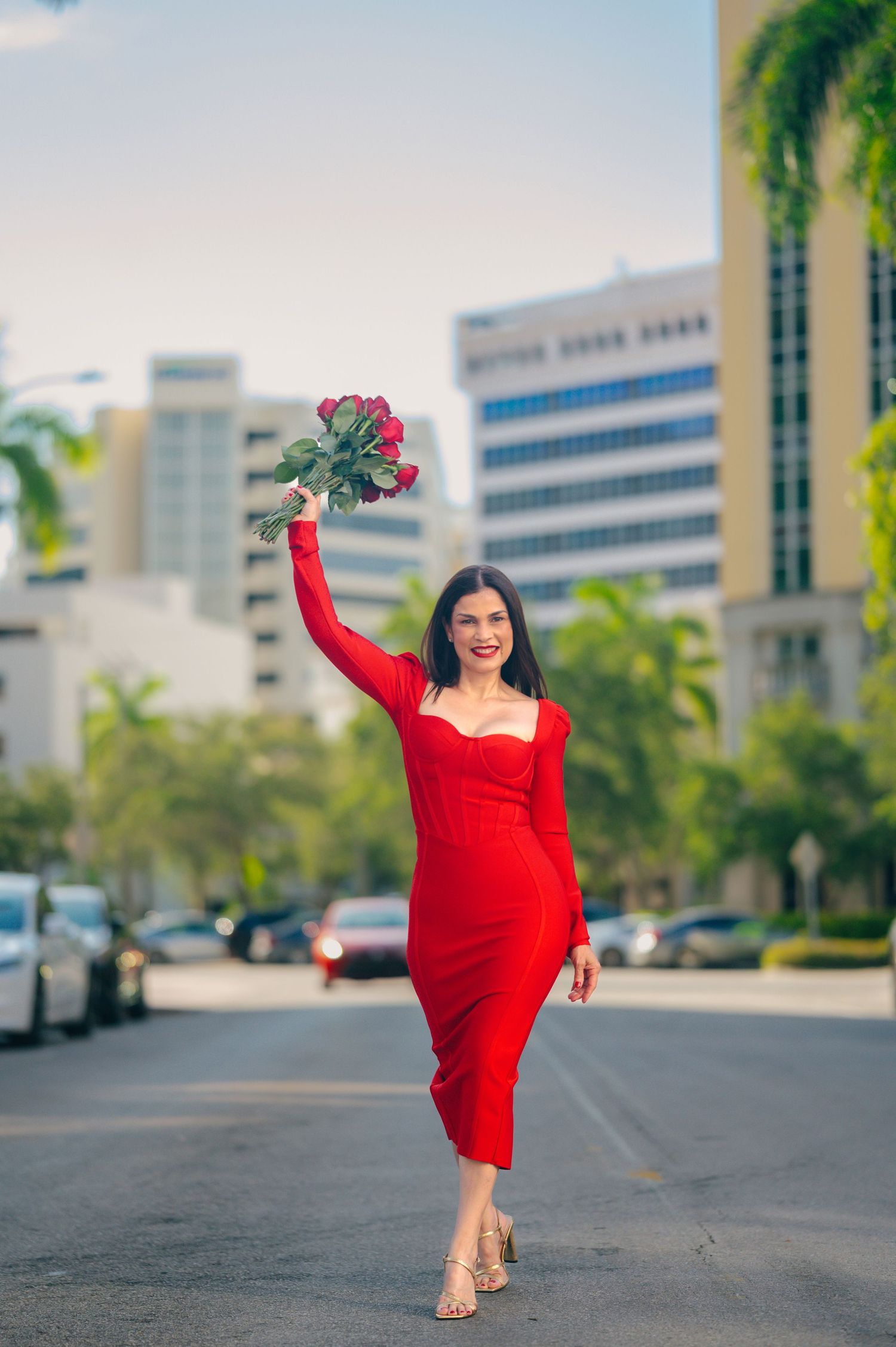 A dancer in a vibrant red dress performs graceful movements in an urban setting with office buildings and palm trees.