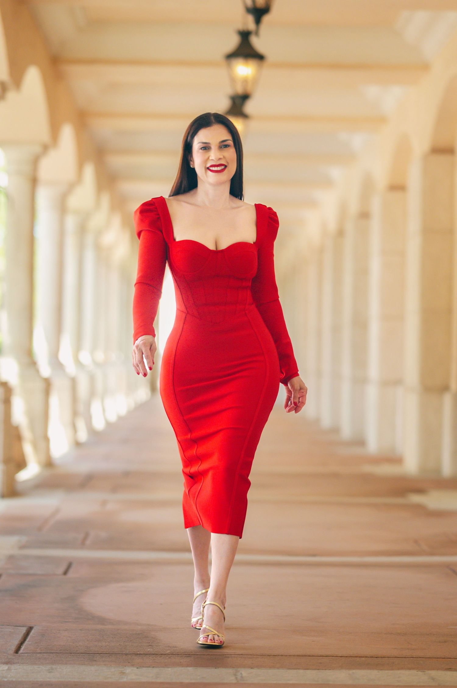 Woman in elegant red dress walks through colonnade hallway with dramatic lighting and architectural columns.