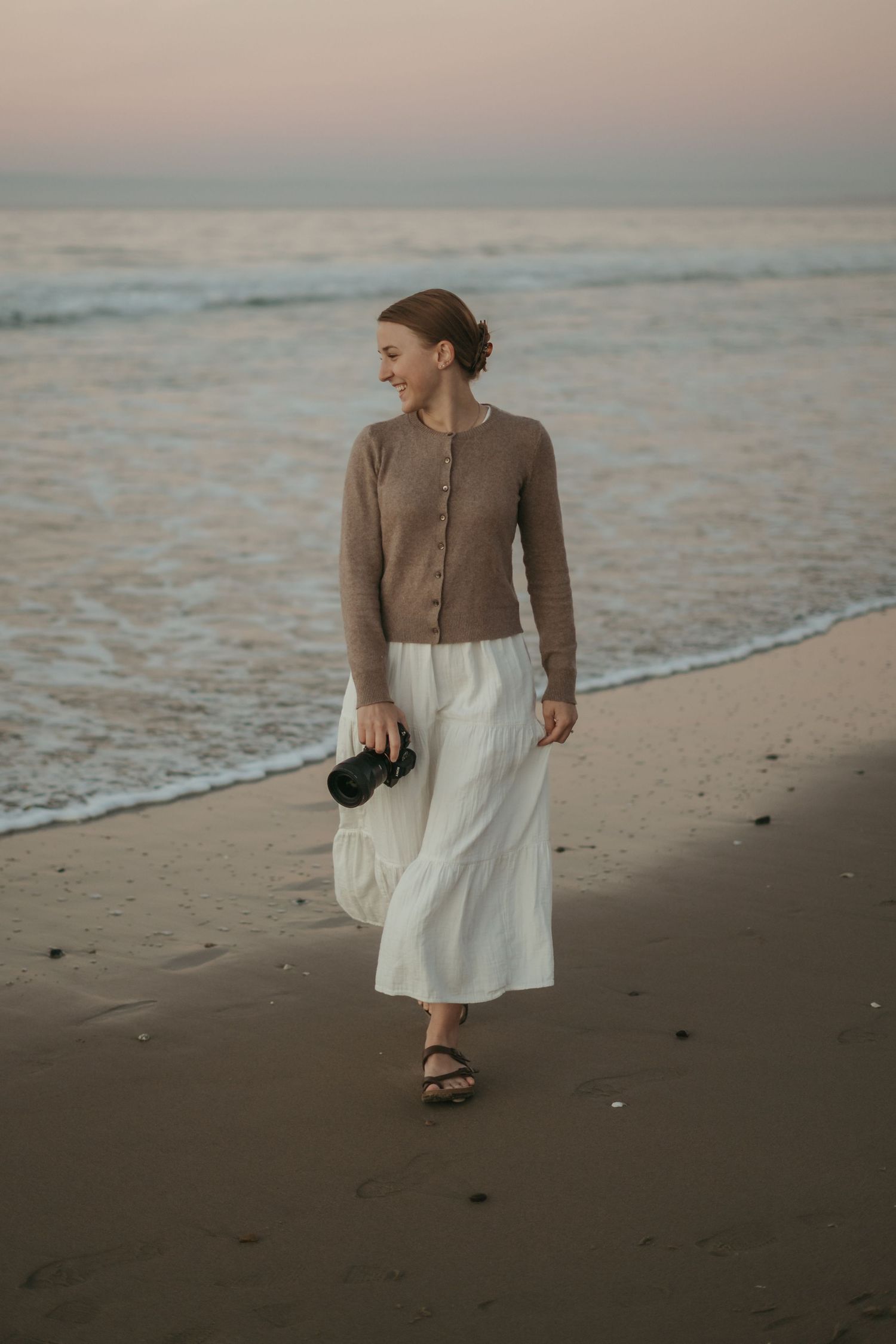 A figure in a brown sweater and white skirt walks along a misty beach shoreline at sunset.
