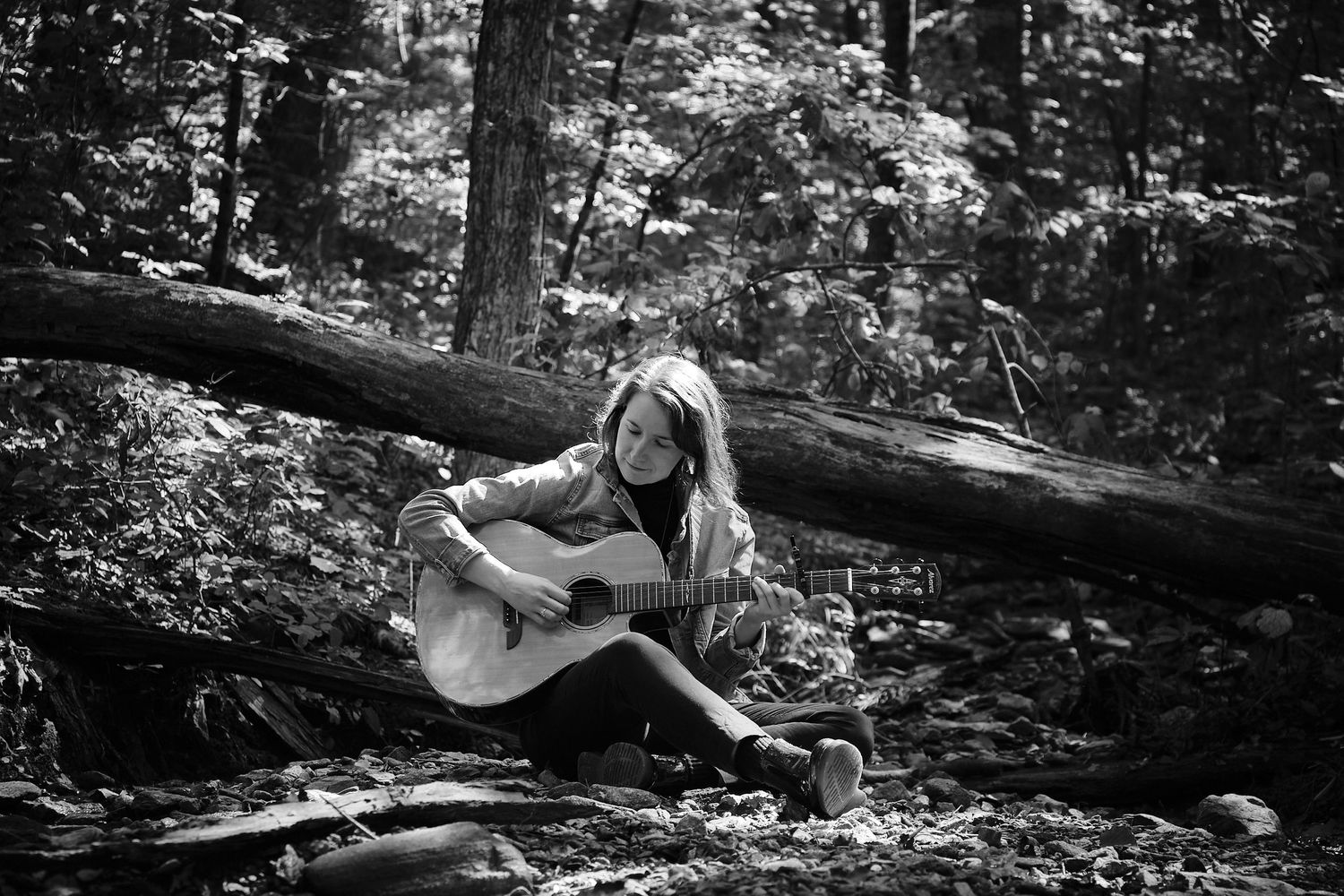 Black and white photo of person playing guitar while sitting on fallen log in forest.