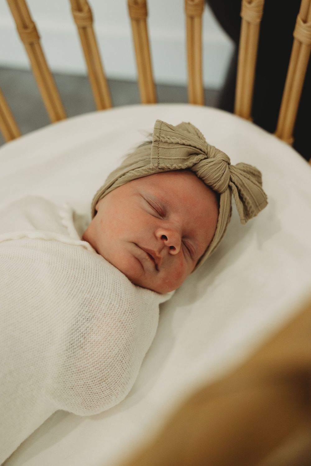 A peaceful sleeping baby swaddled in white blanket with a beige bow headband rests in a wooden crib.