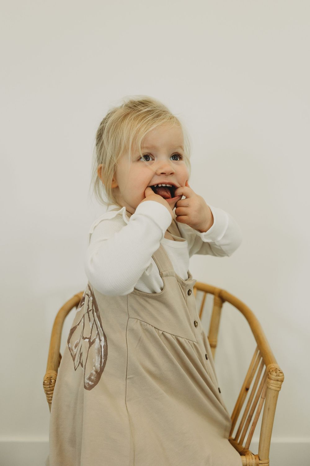 A small child in a beige overall dress and white long sleeve shirt stands by a wooden chair against a light background.