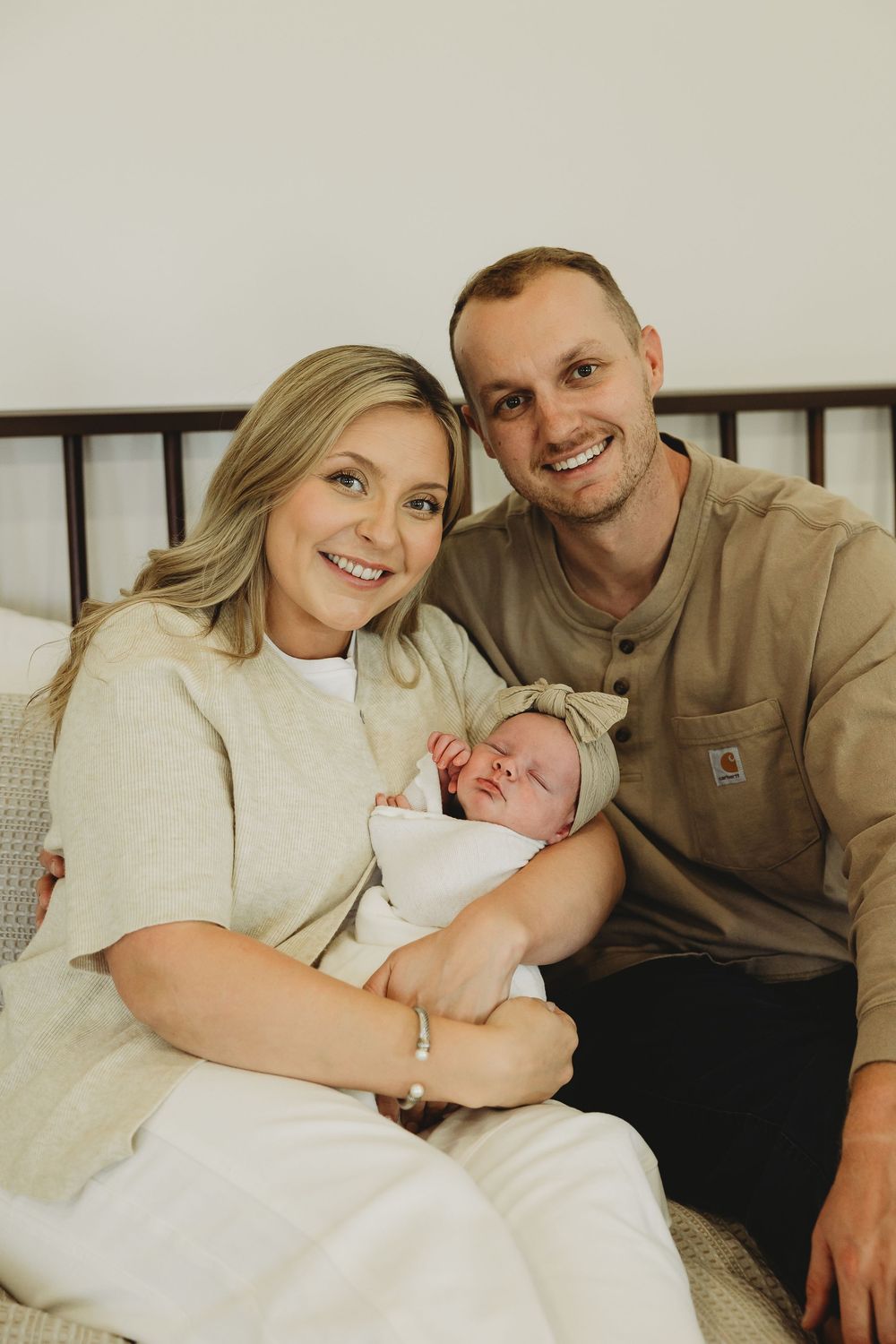 A couple sits together on a couch holding their newborn baby in a cozy home setting.