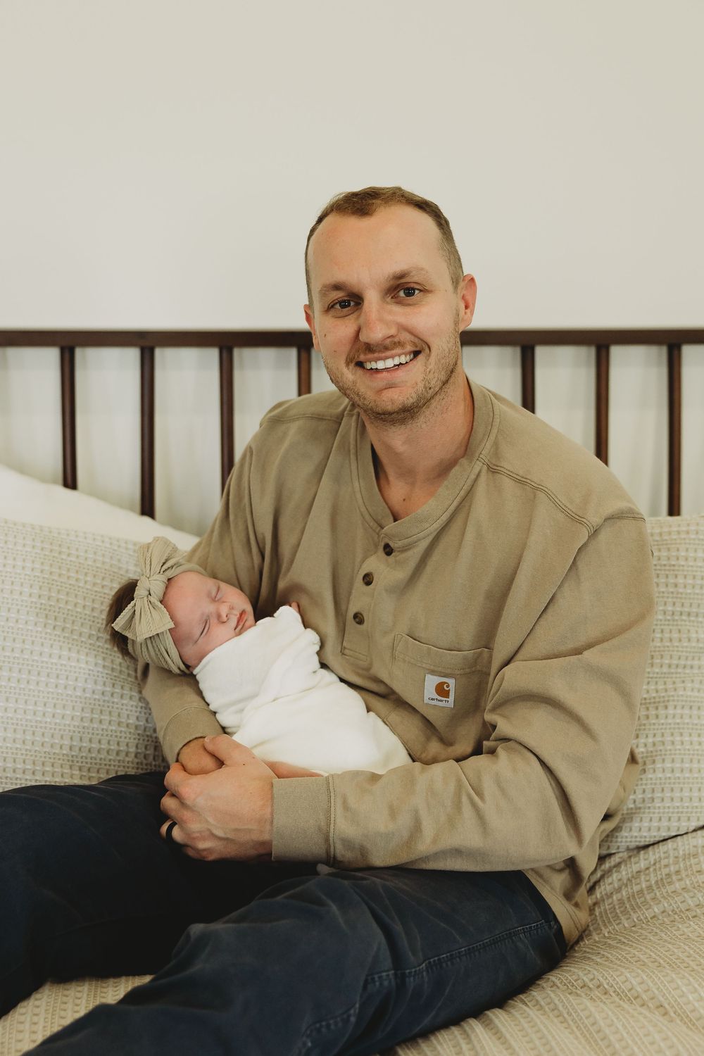 A person wearing a tan Carhartt jacket relaxes on a white couch while holding a sleeping baby in a white outfit.
