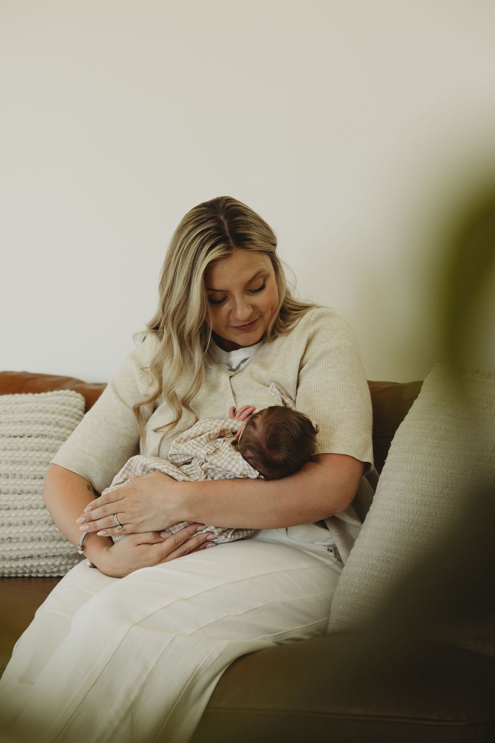 A parent in a cream-colored dress sits on a couch while holding and gazing lovingly at their newborn baby.