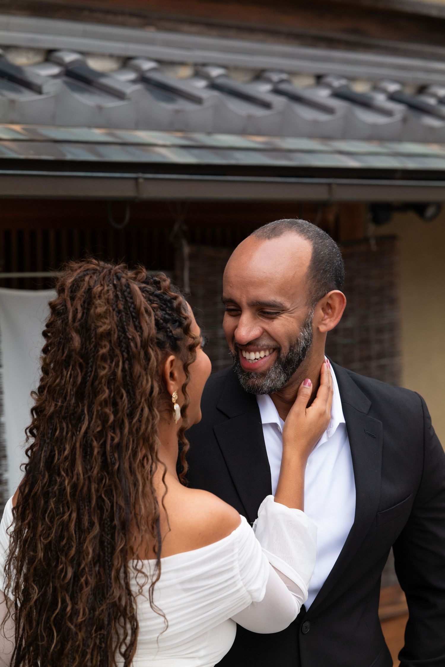 Groom in black suit smiles warmly as bride in white off-shoulder dress tenderly holds his face in Kyoto.