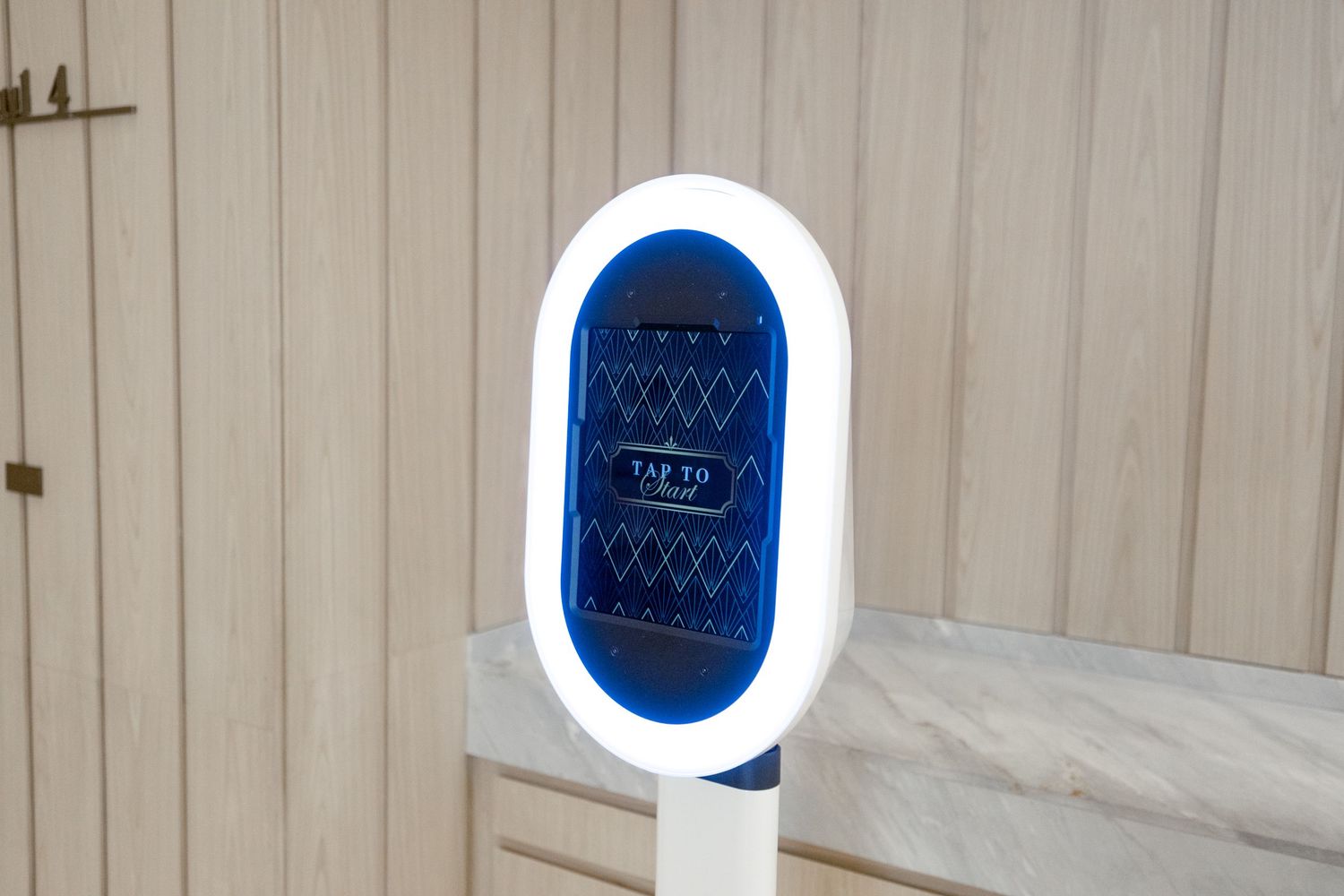 A modern smart mirror display mounted on a beige wall shows a digital interface with geometric patterns glowing in blue light.