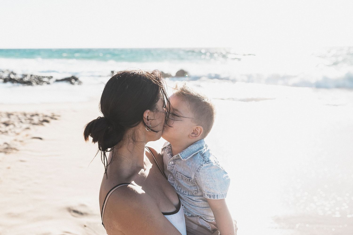 Black and white image showing a mother protecting her daughter from the waves, with a lighthouse on the hedland in the background