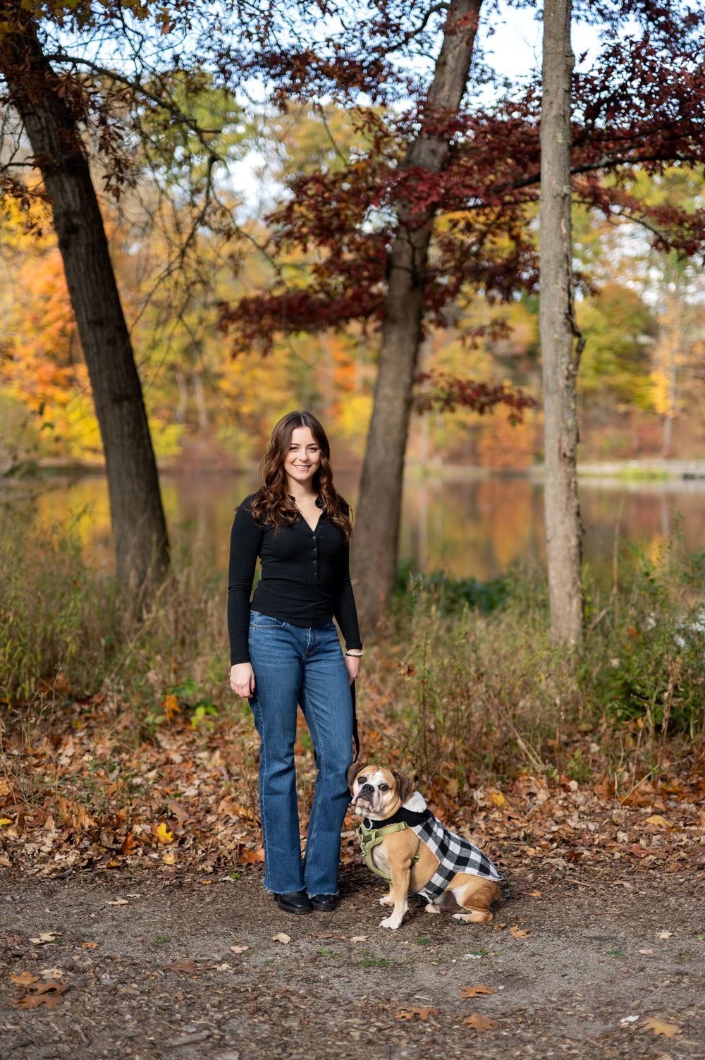 Smiling woman standing with her English Bulldog in a plaid coat in a vibrant autumn park by a lake.