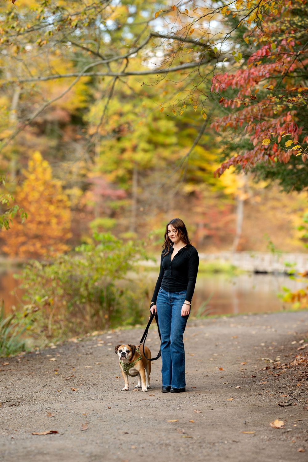 Smiling woman in black top standing with her English Bulldog on a leash on a colorful autumn park trail by a lake.