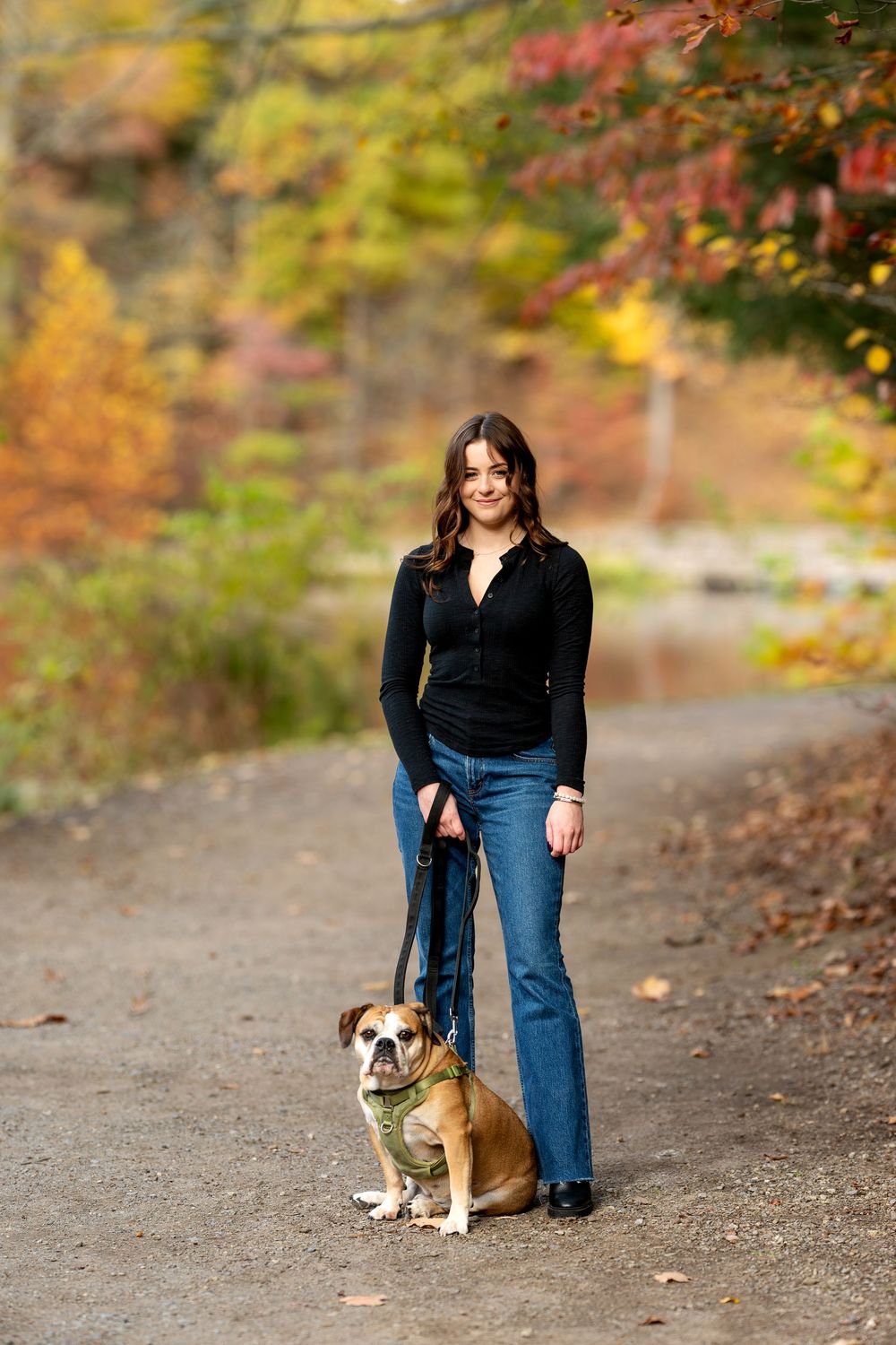 Smiling woman in black top and jeans posing with her English Bulldog on a leash on a scenic autumn park trail.