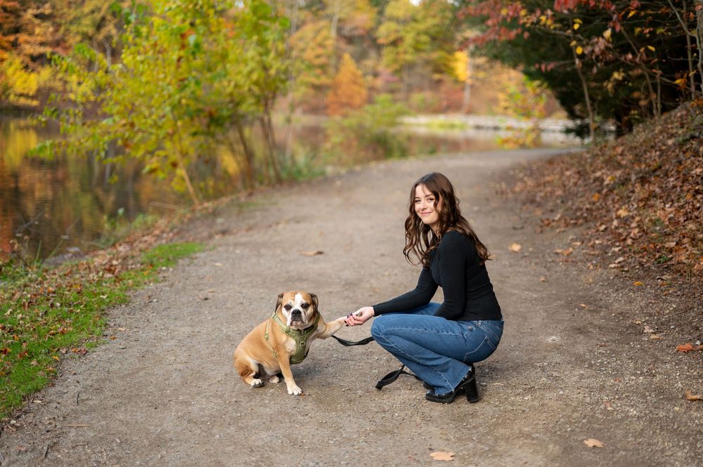 Smiling woman kneeling on a gravel path offering her hand to her English Bulldog amid vibrant autumn foliage in a park.