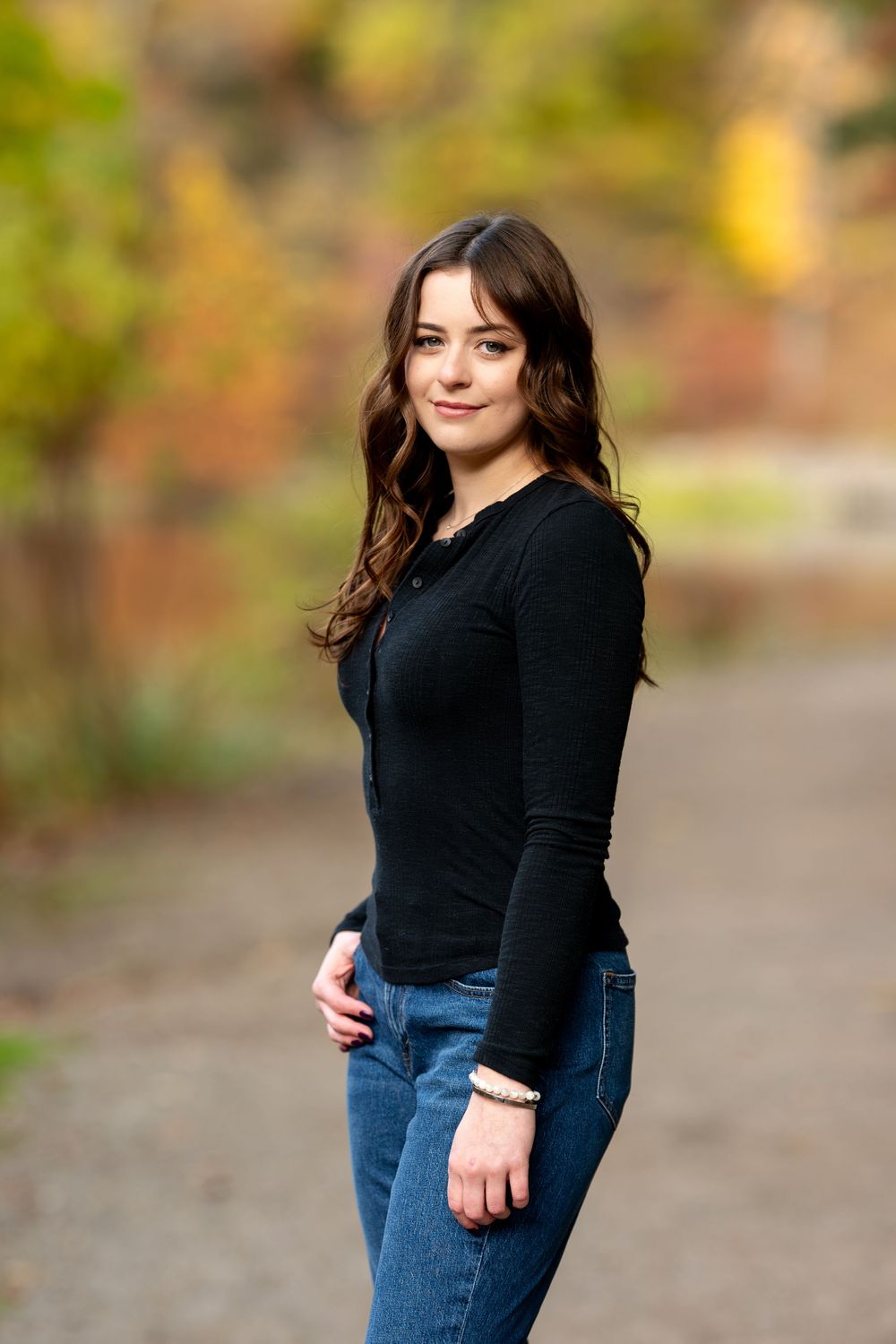 Young woman in black long-sleeve top and blue jeans poses outdoors during autumn.