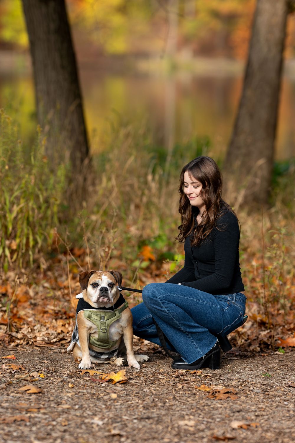 Young woman crouching beside her English Bulldog on a fall forest path with colorful autumn foliage.