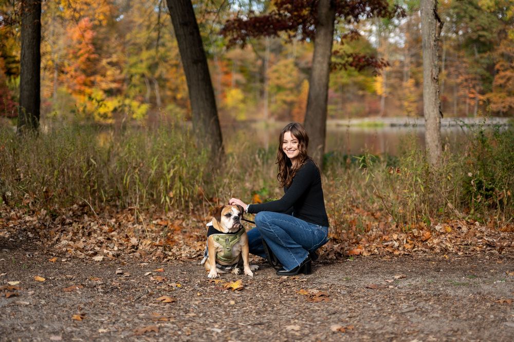 Young woman kneeling beside her English Bulldog on a leaf-covered path in a colorful autumn forest.