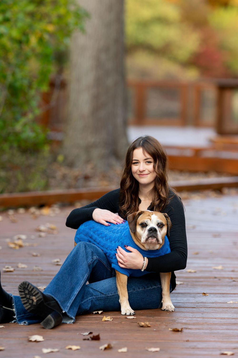 Young woman in black top and jeans sits on wooden boardwalk hugging her English Bulldog wearing a blue sweater in autumn park.