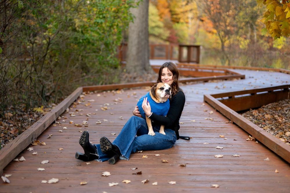Woman sitting on fall foliage-covered boardwalk cuddling her English Bulldog in a blue outfit among colorful autumn trees.