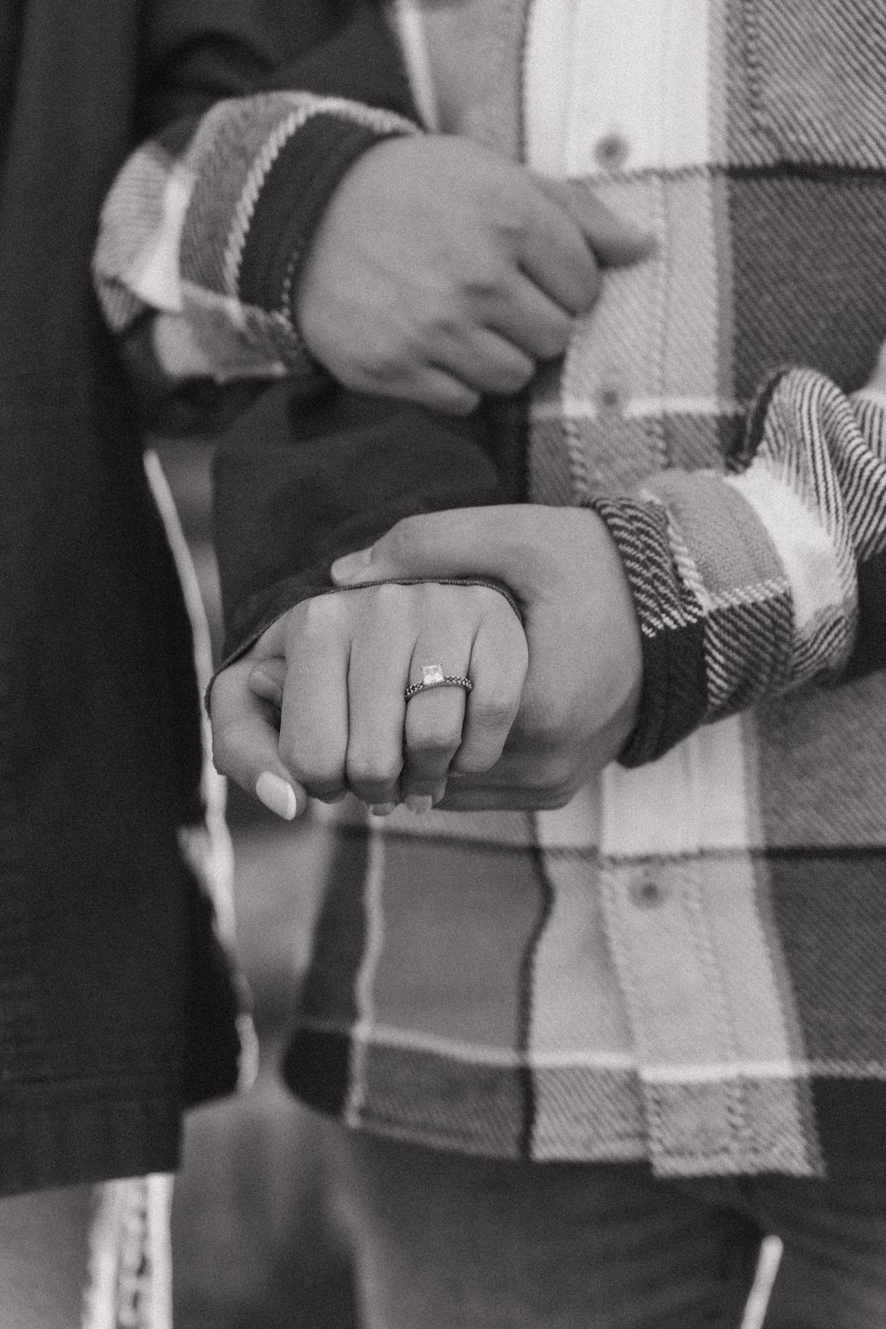 Black and white close-up of clasped hands wearing a plaid shirt sleeve, with an engagement ring visible.