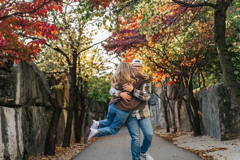 A couple embraces while walking along a tree-lined path with vibrant red and orange fall foliage overhead.