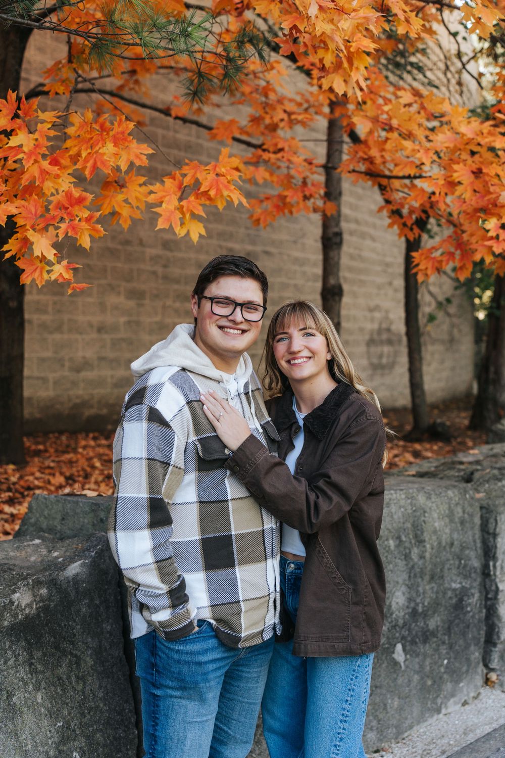A couple poses together against a brick wall with vibrant orange autumn leaves overhead.