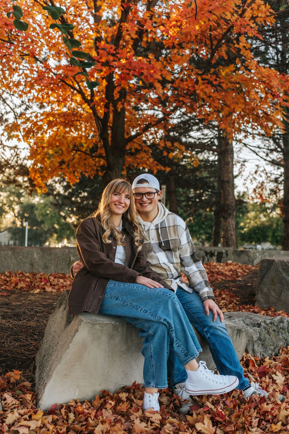 A couple sits together on a stone wall surrounded by vibrant orange autumn foliage in a romantic outdoor setting.