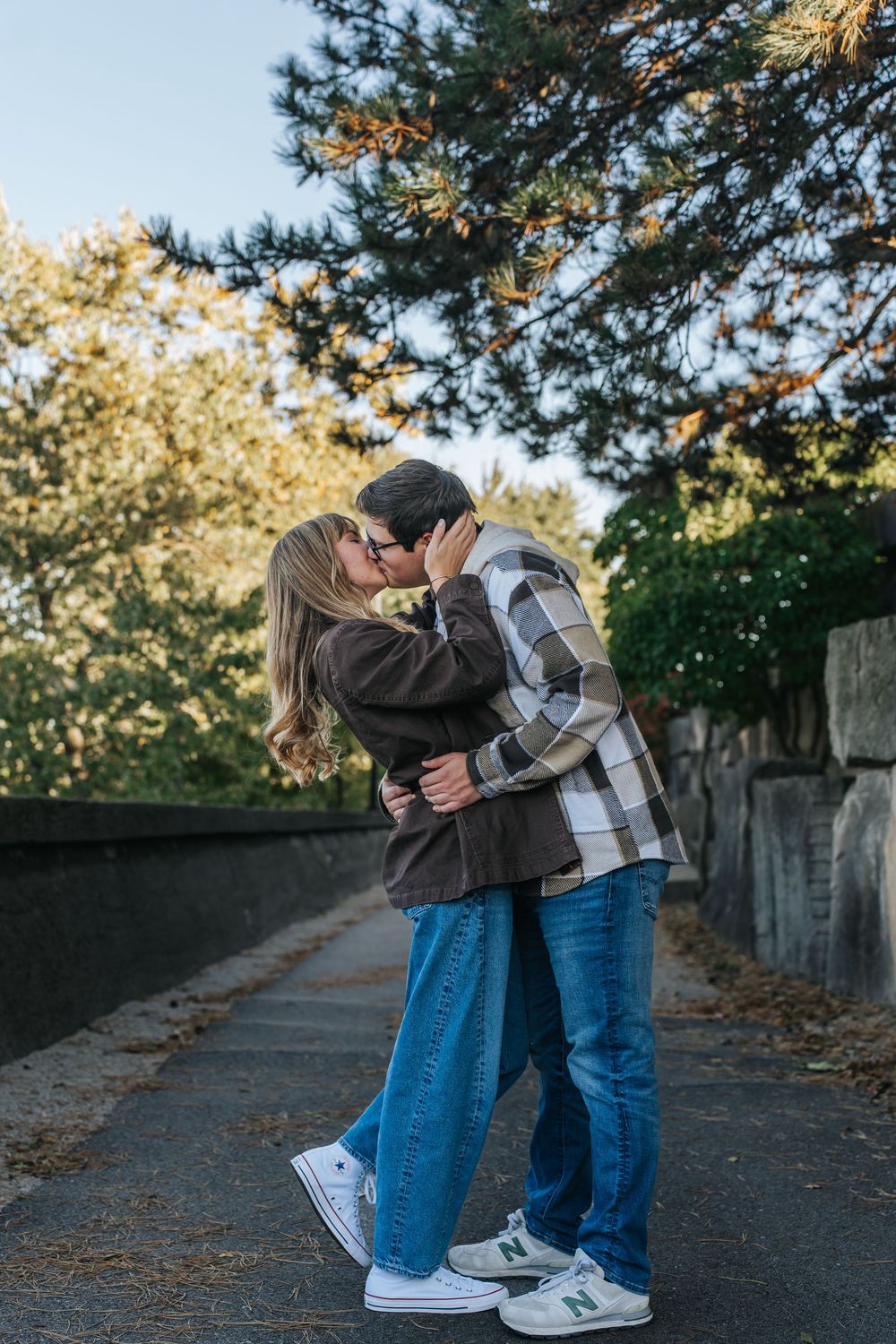 A couple embraces on a concrete path with pine trees and autumn foliage in the background.