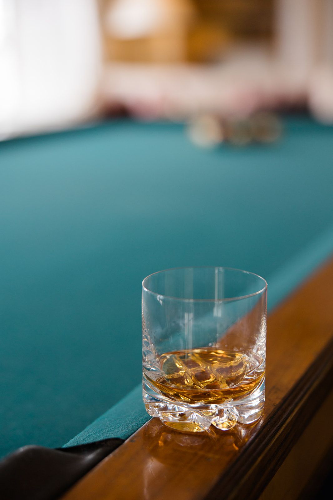 A glass of whiskey with ice sits on a dark surface with a teal-colored background creating a moody atmosphere.
