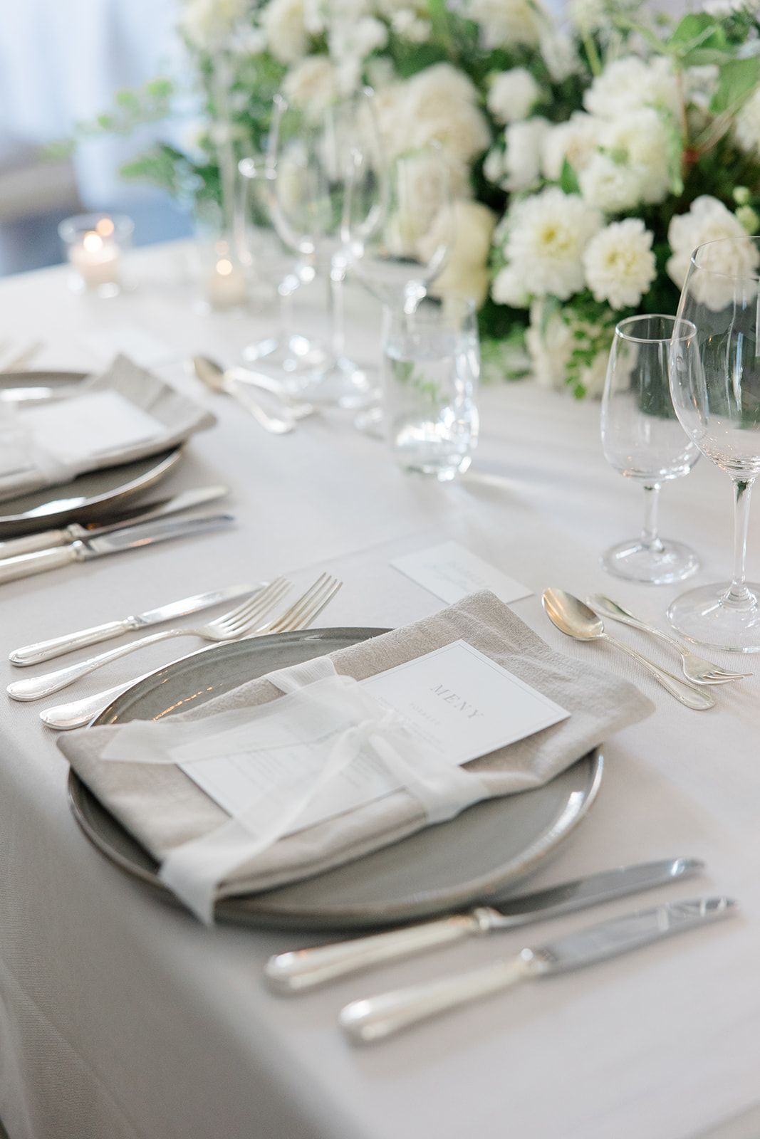 Elegant table setting with white napkins, silver cutlery and delicate white floral arrangements on a crisp tablecloth.
