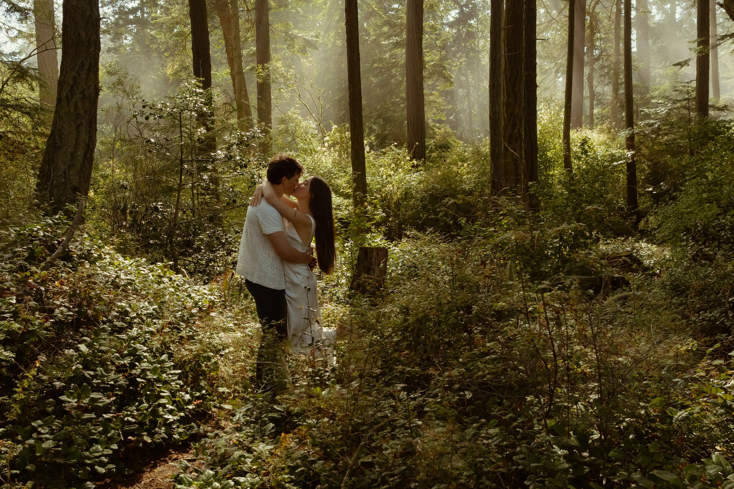 A couple embrace in a misty green forest with sunlight filtering through the trees captured by Savvy Photography - Seattle Wedding Photo