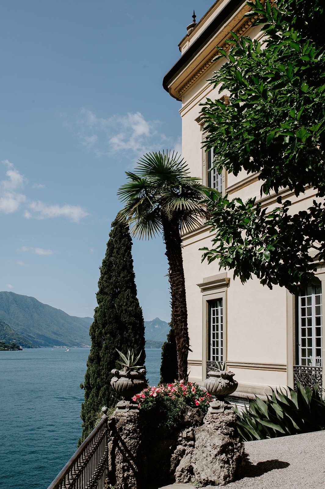 Elegant villa with cypress trees and palm fronds overlooking Lake Como's blue waters and mountain vistas.