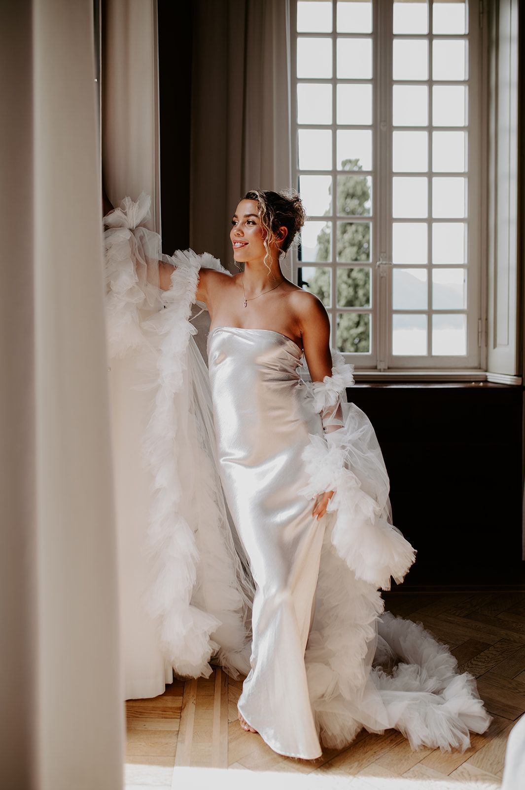 Artistic motion shot of flowing white wedding dress fabric in a sunlit room with French windows.