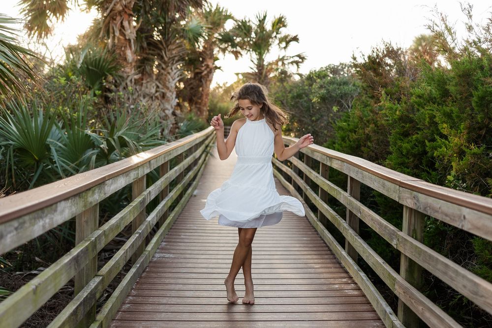 A young girl in a white dress twirls on a wooden boardwalk surrounded by palm trees at sunset.