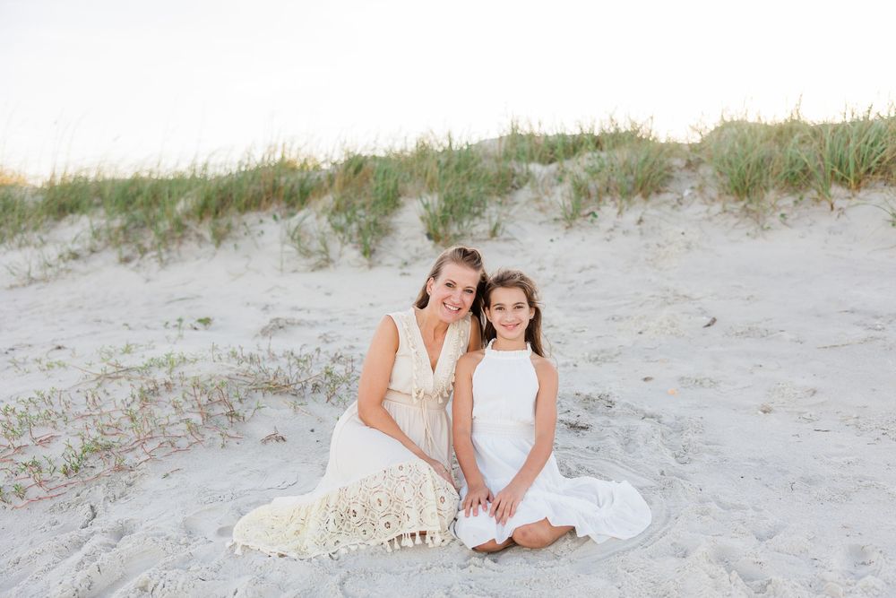 A mother and daughter in white dresses sit together on sandy beach dunes.