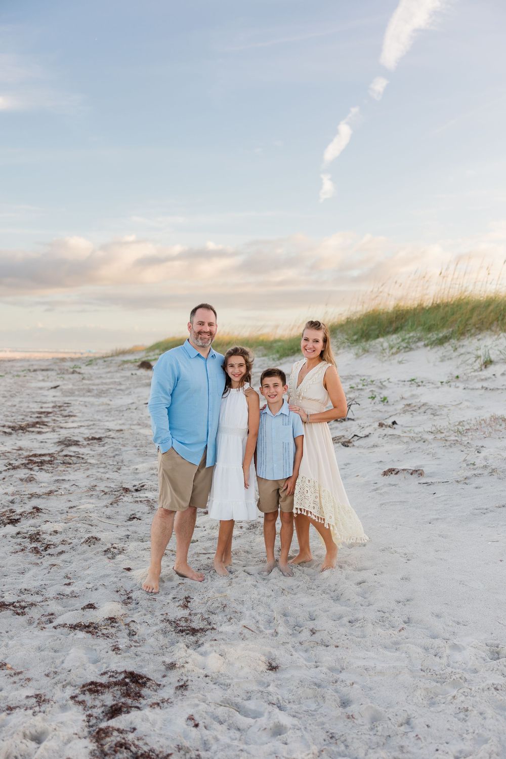A family dressed in light blue and white poses together on a sandy beach at sunset.