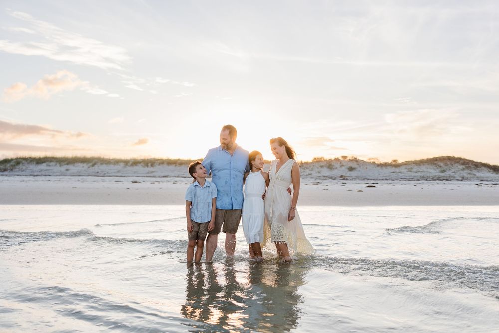 Family standing together in shallow ocean water during sunset.